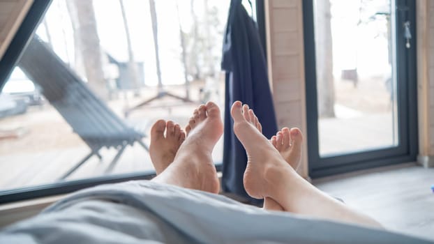 The feet of a married couple sticking out from under the blanket in a country house with panoramic windows