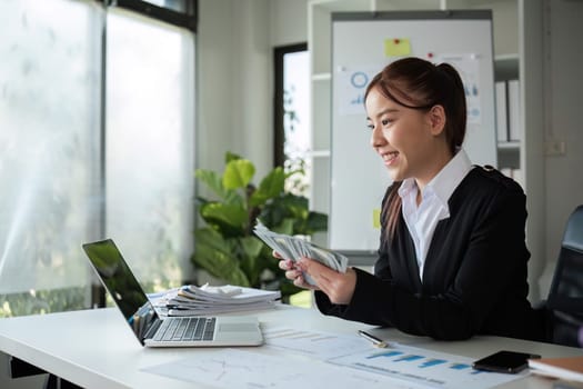 Successful Asian businesswoman counting money and checking stock market data on laptop in office.