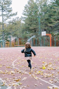 Little girl runs along a basketball court strewn with fallen leaves in the park. Back view. High quality photo