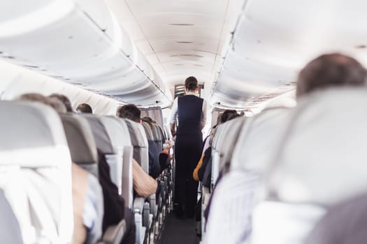 Interior of airplane with passengers on seats and stewardess in uniform walking the aisle, serving people. Commercial economy flight service concept