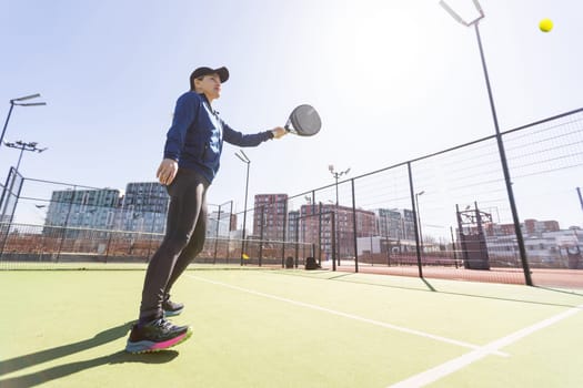 A girl in sportswear is training on a paddle tennis court. The girl is hitting the ball against the glass to make a rebound. Concept of women playing paddle. High quality photo