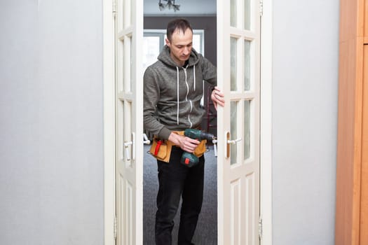 Maintenance Concept. Focused serious young locksmith with tattoo on hand wearing blue uniform standing on one knee in doorway installing doorknob handle, repairing wooden front door entrance. High quality photo