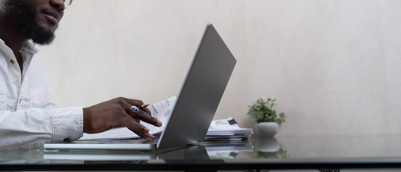 African American man working with laptop computer and using calculator, making financial audit, reviewing bills tax and accounting in living room. Black guy do freelance work at home office.