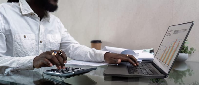 African American man working with laptop computer and using calculator, making financial audit, reviewing bills tax and accounting in living room. Black guy do freelance work at home office.