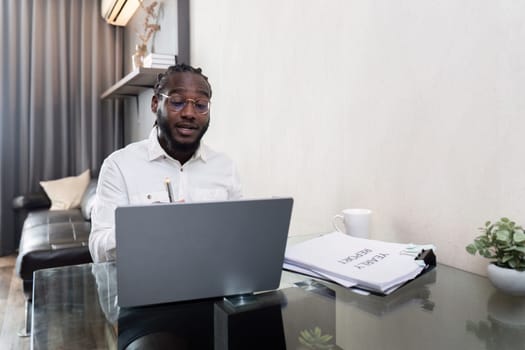 African American man working with laptop computer remote while sitting at glass table in living room. Black guy do freelance work at home office.