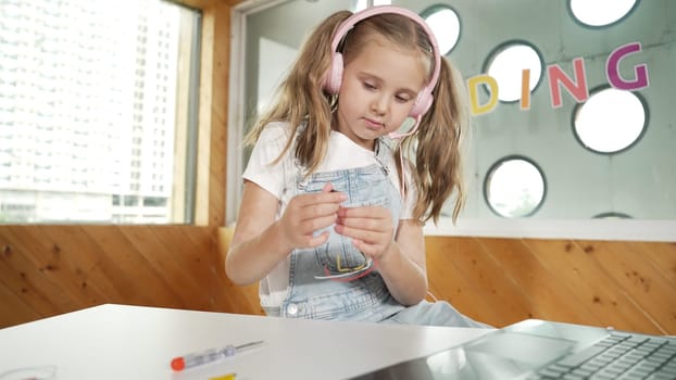 Pretty girl wearing headphone while study electronic equipment. Caucasian child doing science experiment while laptop, screwdriver and wires placed near on table. Smart online classroom. Erudition.