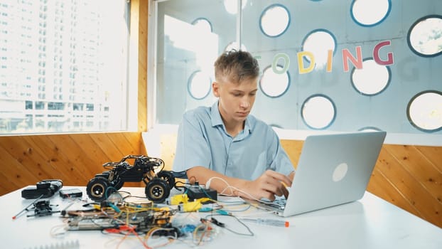 Smart student working on laptop while analysis car model construction. Happy highschool boy using computer to study or learn about robotic model structure on table with electronic tool. Edification.