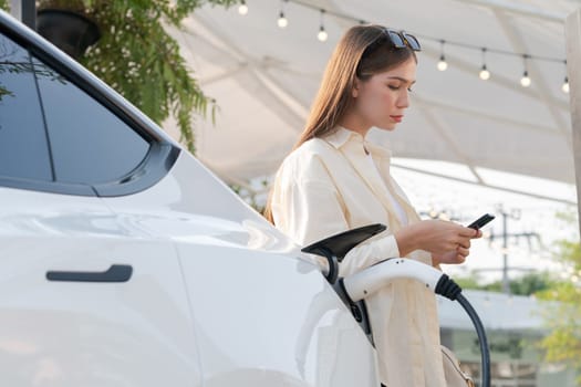 Sustainable urban commute with young woman pay electricity for EV electric car recharging at outdoor cafe in springtime garden, green city sustainability and environmental friendly EV car. Expedient