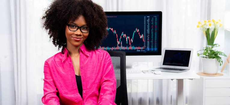 Analytical young African American businesswoman, a specialist in successful stock exchange trading, against dynamic data graph displaying marketing trend analysis on screen. Tastemaker.