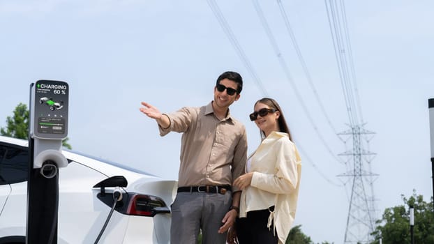 Young couple recharge EV car battery at charging station connected to power grid tower electrical industrial facility as electrical industry for eco friendly vehicle utilization. Expedient