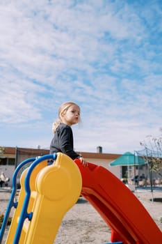 Little girl sits on a colorful slide at the playground and looks away. High quality photo