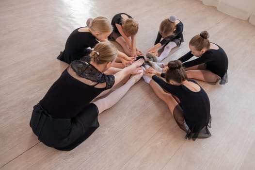 Caucasian woman and five little girls sit in a circle and do stretching at a ballet school