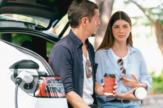 Lovely young couple drinking coffee while recharging battery for electric car during road trip travel EV car in natural forest or national park. Eco friendly travel during vacation and holiday. Exalt