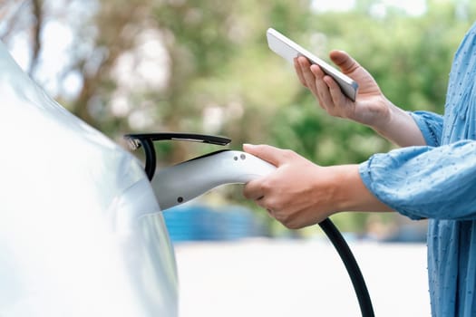 Hand insert EV charger plug into electric vehicle to recharge EV car, battery status display on smartphone EV application. Future alternative clean and sustainable energy for transportation. Perpetual