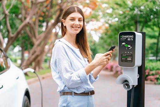 Young woman using smartphone online banking application to pay for electric car battery charging from EV charging station during autumn vacation holiday trip at national park or autumnal forest. Exalt