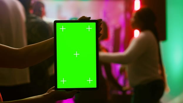 Girl showing greenscreen display at club holding blank chroma key display at party on dance floor. Modern person using isolated mockup screen with copy space template on device. Tripod shot.