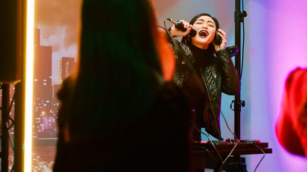 Asian woman mixing music at DJ station, creating funky club atmosphere for group of people on dance floor. Young cheerful adult using audio stereo equipment at nightclub, partying.