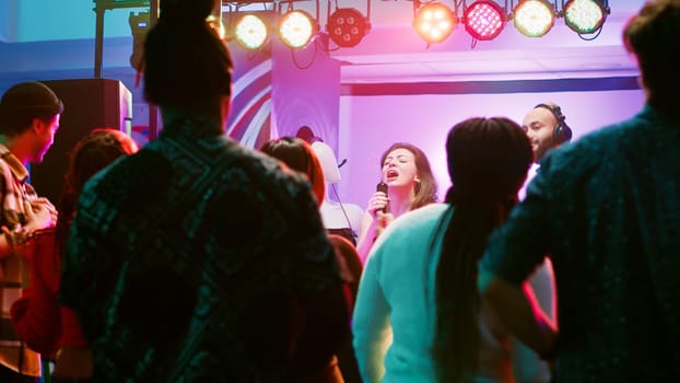 Happy woman doing karaoke at party, singing songs with DJ in front of friends on dance floor. Confident girl having fun with live show perfromance next to electronic mixing station at nightclub.