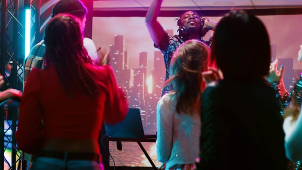 Young man clubbing with people at bar, having fun with DJ mixing station on nightclub stage with disco ball. Funky person creating live performance on audio panel, spotlights at club.