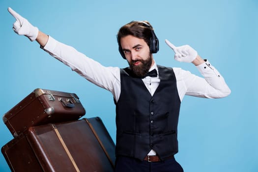 Blue background with enthusiastic bellboy singing and enjoying tunes while he is using audio earpieces. Person in a suit dancing and behaving openly at the camera, having fun with music.