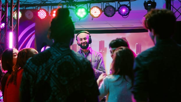 Male DJ putting music at club party, mixing electronic sounds at audio equipment station. Diverse group of friends jumping around and dancing on dance floor, clubbing entertainment.