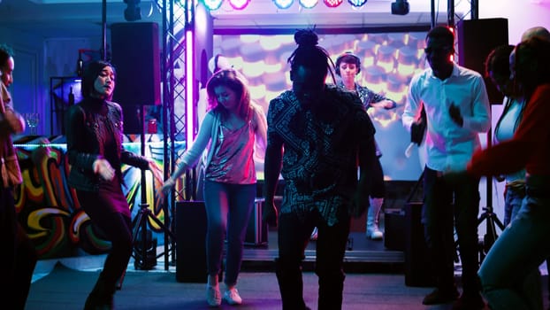 Happy persons jumping at dance party, having fun at social gathering in discotheque. Young funky friends showing dance moves on music at club, colorful spotlights. Handheld shot.