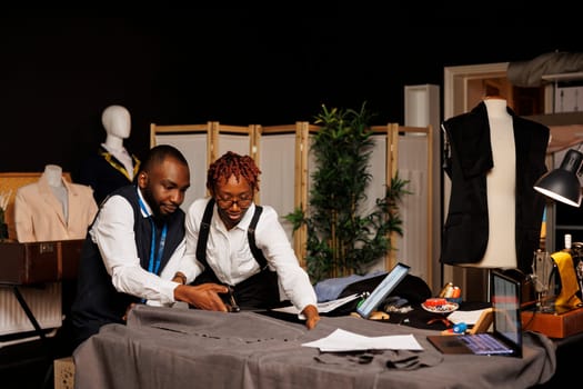 Well dressed couturiers cutting premium fabric in tailoring studio during hand stitched suitmaking process. African american fashion designers preparing textile material for bespoke clothing