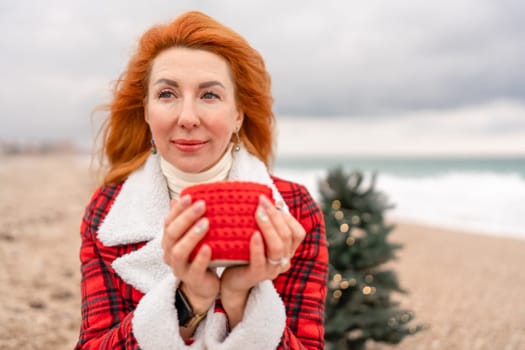 Lady in plaid shirt with a red mug in her hands enjoys beach with Christmas tree. Coastal area. Christmas, New Year holidays concep.