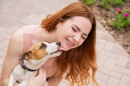 Dog jack russell terrier licks the owner in the face outdoors. Girl with braces on her teeth