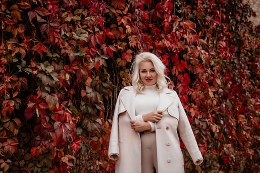 A blonde woman is standing in front of a wall covered in red leaves. She is wearing a white coat and holding her arms out to the side. Concept of warmth and comfort