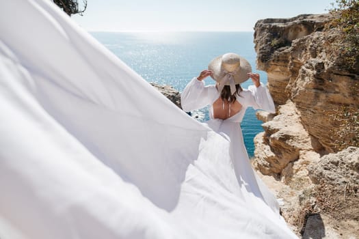 A woman in a white dress is standing on a rocky cliff overlooking the ocean. She is wearing a straw hat and she is enjoying the view