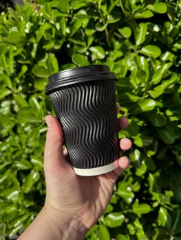 Hand holding black corrugated cardboard coffee cup with lid against background of lush green leafy bush. Urban eco friendly lifestyle. Blank space for sticker or label. High quality photo