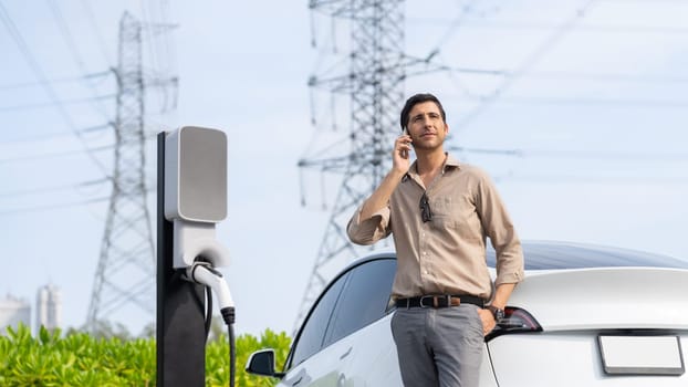 Man talking on the phone while recharge EV car battery at charging station connected to power grid tower electrical as electrical industry for eco friendly car utilization. Panorama Expedient