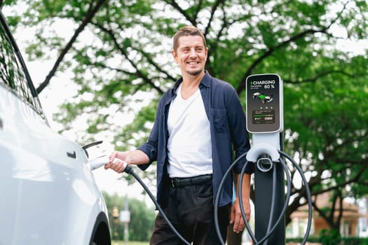 Man recharge EV electric vehicle's battery from EV charging station in outdoor green city park scenic. Eco friendly urban transport and commute with eco friendly EV car travel. Exalt