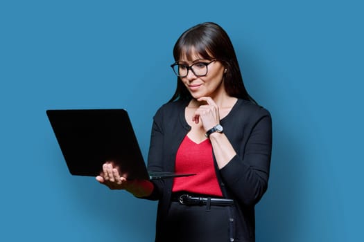 Smiling business middle aged woman using laptop on blue studio background. Confident successful mature female holding computer in hands. Business, work, job, lifestyle, technology, 40s people concept