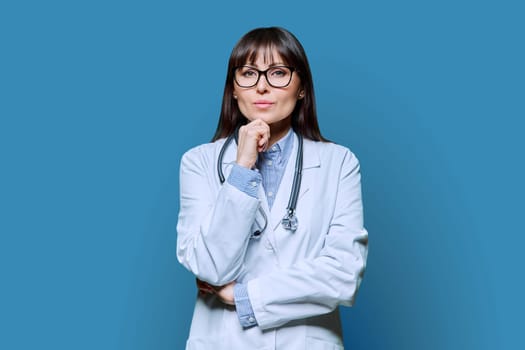 Confident serious middle-aged female doctor in white lab coat with stethoscope, looking at camera on blue studio background. Healthcare, medicine, staff, treatment, medical services concept