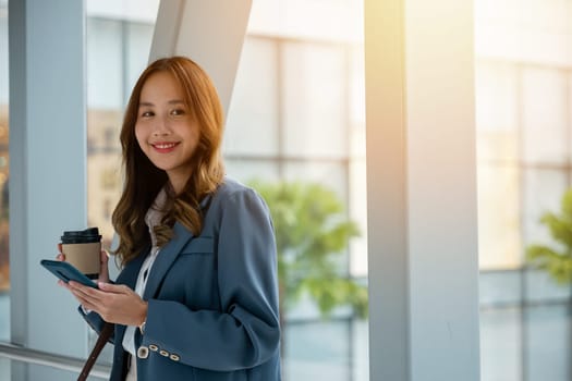 Asian woman on escalator holding a hot cup of coffee and smartphone. Perfect for lifestyle and communication-related themes. City road and building background. Business lifestyle on the go concept