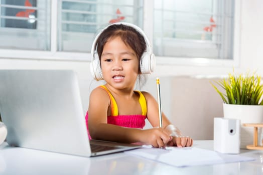 Asian child girl studying video conference distant education at home. Little kid preschool wear headphones sit at desk use laptop computer and communicates on Internet online video call with teacher