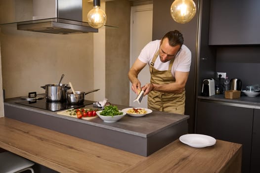 Male chef cooking in the home kitchen, grating parmesan cheese over a plate with freshly boiled pasta with tomato sauce. Ingredients on the table in stylish modern minimalist home kitchen interior