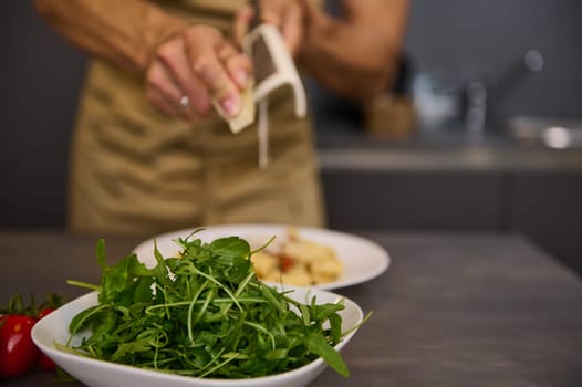 Closeup male chef cooking dinner at home, grating parmesan cheese on a pasta with tomato sauce. A bowl with fresh clean arugula leaves on the foreground. Food. Culinary. Italian cuisine