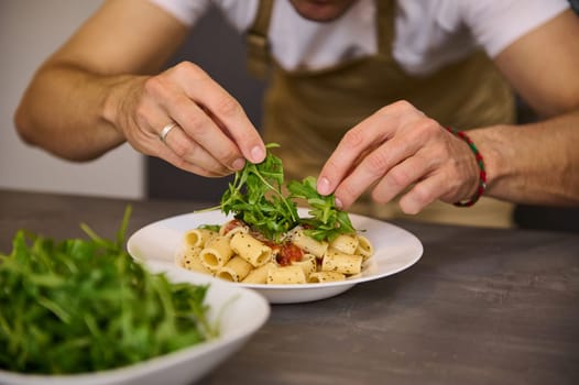 Close-up male chef hands garnishing Italian pasta with fresh green arugula leaves. Man cooking pasta according to traditional Italian recipe. Food background. Selective focus