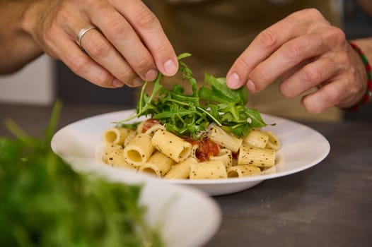 Close-up hands of a chef in home Kitchen, preparing and decorating meal with fresh arugula leaves, preparing pasta with tomato sauce according to traditional Italian recipe. Culinary. Cuisine. Epicure