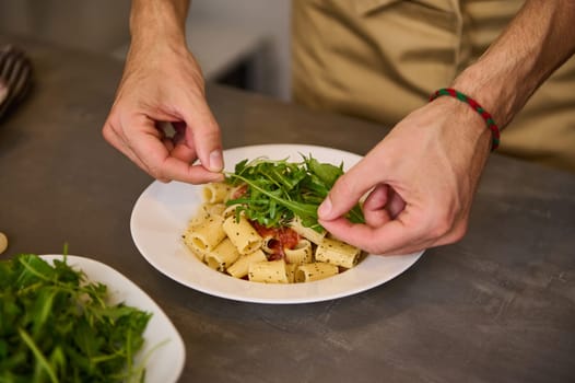 Close-up view from above hands of a chef preparing and decorating meal with fresh arugula leaves, preparing pasta with tomato sauce according to traditional Italian recipe. Cuisine. Culinary. Epicure