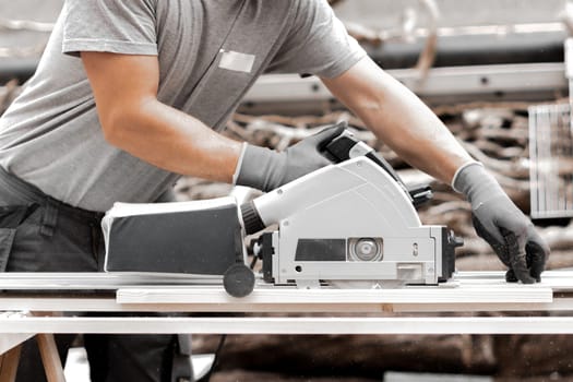 One young Caucasian unrecognizable man in a uniform and gray gloves is sawing a board into beams with an electric saw, standing in the backyard of a house on a summer day, close-up side view.