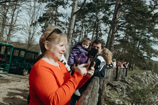 Portrait of one beautiful Caucasian elderly woman in a red sweater with a satisfied emotion and a smile on her face photographing on a smartphone a panoramic view from the mountain of a nature reserve in Rochefort, Belgium, on a spring sunny day, close-up side view.
