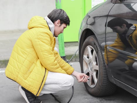 Young caucasian man in a yellow jacket and gray sweatpants, squatting, spins the bearing with his fingers into the wheel of a car for pumping air in a parking lot, side view with selective focus and closeup.Gas station concept.