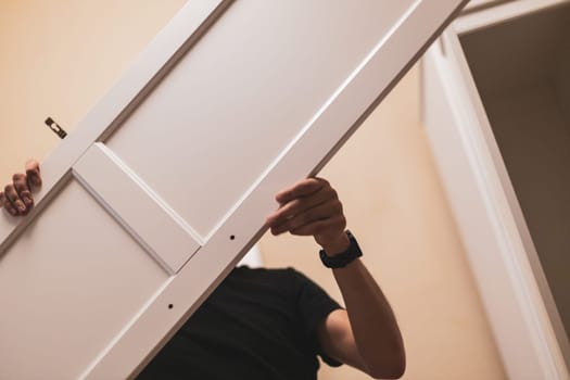 Young caucasian brunette man holding with hands the side door of the wardrobe to install inside, standing in the bedroom, close-up view from below with selective focus. Furniture assembly concept.