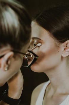 One young handsome Caucasian makeup artist applies eye shadow with a brush to the left eyelid of a girl sitting sideways in a chair early in the morning in a beauty salon, close-up side view with depth of field. Step by step.