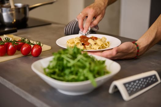 Close-up of chef's hands using kitchen towel, wiping away traces of tomato sauce on a plate of freshly prepared Italian pasta. Fresh green arugula leaves and kitchen utensils on blurred background