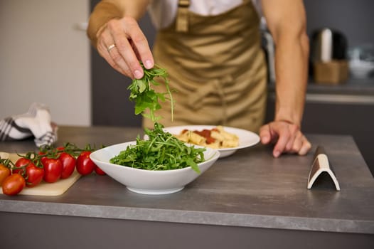 Close-up chef hands holding fresh arugula leaves, garnishing meal, cooking Italian spaghetti in the home kitchen. Selective focus
